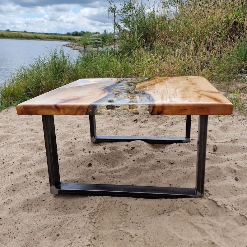 K&ouml;nnte beinhalten: Ein Holz-Couchtisch mit einem durchsichtigen Epoxidharz-Fluss, der durch die Mitte verl&auml;uft. Der Tisch hat ein schwarzes Metallgestell und steht auf einer sandigen Oberfl&auml;che.