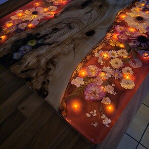 May include: A live edge wood table featuring a central resin river filled with pressed flowers and embedded warm LED lights. The resin has a reddish-pink tint, and the wood displays natural grain patterns, knots, and dark brown streaks throughout.