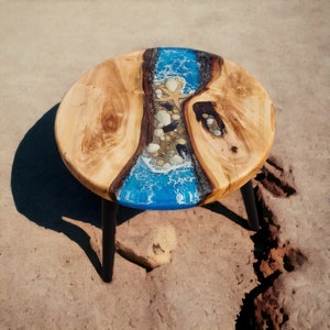 Puede incluir: Una mesa redonda de madera con un diseño de río de epoxi azul y conchas incrustadas. La mesa tiene patas de metal negro.
