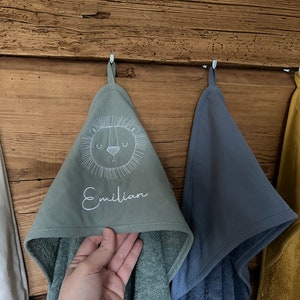 May include: Three hooded towels hanging on hooks. The towel in the center is green with a white embroidered lion face and the name "Emilian". The towel on the left is white and the towel on the right is yellow.