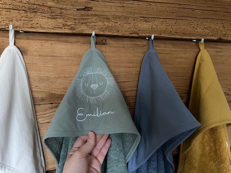 May include: Three hooded towels hanging on hooks. The towel in the center is green with a white embroidered lion face and the name "Emilian". The towel on the left is white and the towel on the right is yellow.