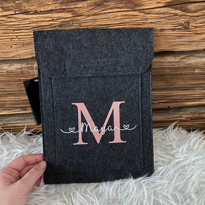 Puede incluir: Una funda para portátil de fieltro gris con una letra M de fieltro rosa y el nombre Maya escrito en cursiva encima.