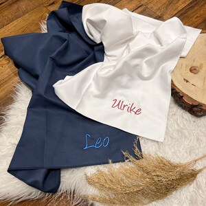 May include: Two white fabric towels with blue and red embroidery. The towels are folded and have the names "Leo" and "Ulrike" embroidered on them.