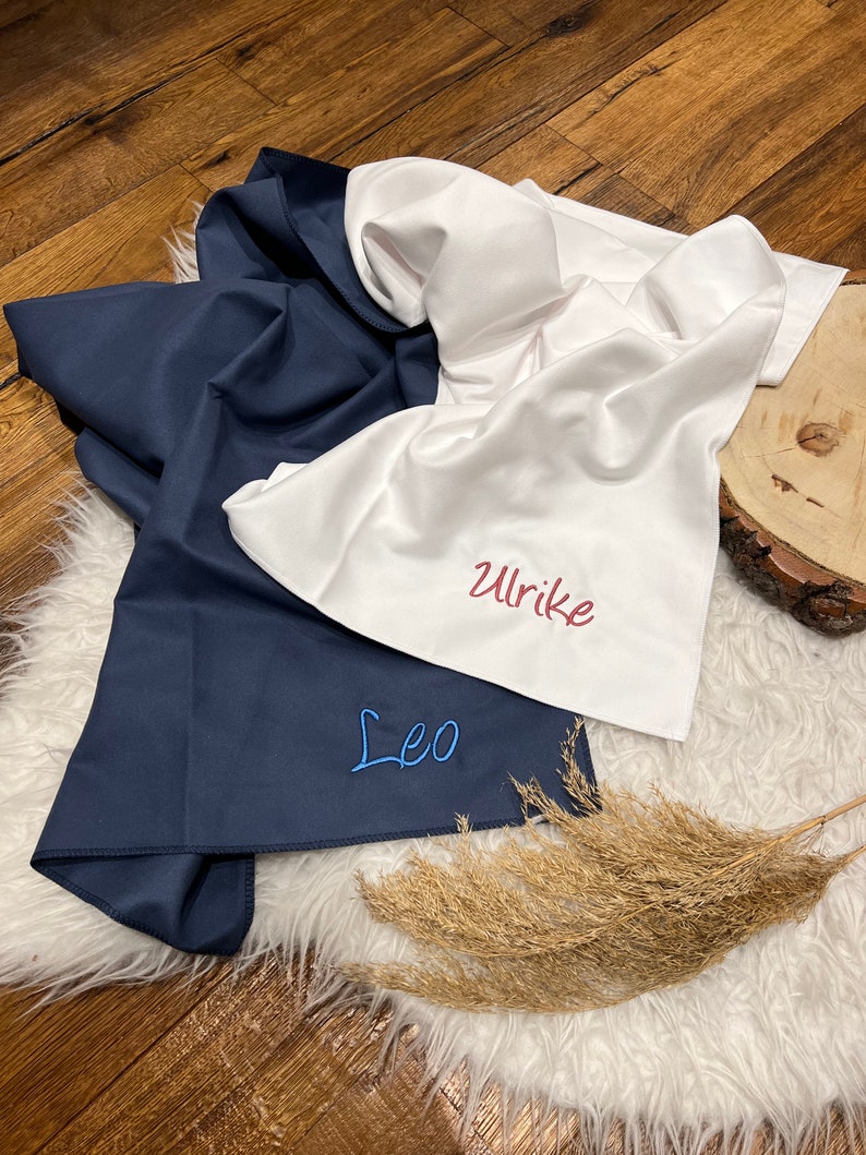 May include: Two white fabric towels with blue and red embroidery. The towels are folded and have the names "Leo" and "Ulrike" embroidered on them.