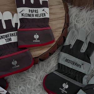 Może przedstawiać: Szare, białe i czerwone rękawice robocze z napisem "PAPAS KLEINER HELFER" i "STRAUSS". Rękawice mają czerwone wykończenie i białe sekcje na palce. Na rękawicach nadrukowano r&oacute;wnież klucz i młotek.