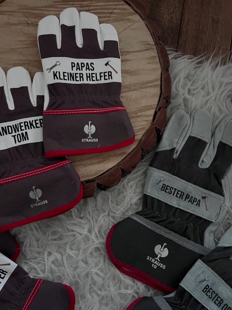 Może przedstawiać: Szare, białe i czerwone rękawice robocze z napisem "PAPAS KLEINER HELFER" i "STRAUSS". Rękawice mają czerwone wykończenie i białe sekcje na palce. Na rękawicach nadrukowano r&oacute;wnież klucz i młotek.
