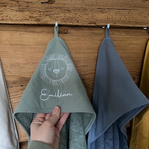 May include: Four hooded towels hanging on hooks. The towel in the center is green with a white embroidered lion face and the name "Emilian". The towel to the right is blue, the towel to the left is white, and the towel on the far left is yellow.