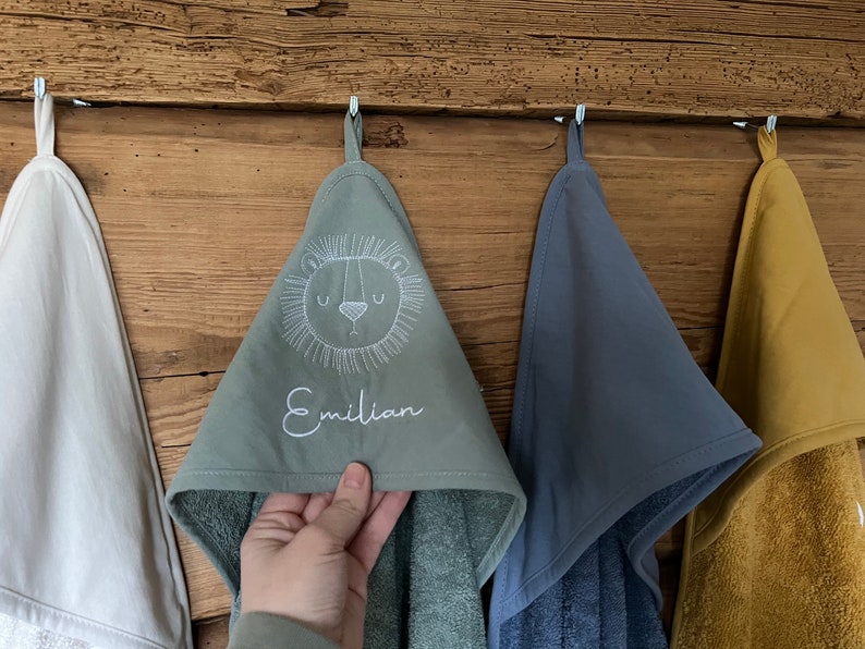 May include: Four hooded towels hanging on hooks. The towel in the center is green with a white embroidered lion face and the name "Emilian". The towel to the right is blue, the towel to the left is white, and the towel on the far left is yellow.
