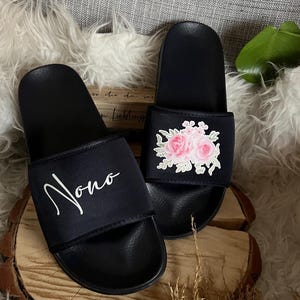 May include: A pair of black slides with a floral design on one slide and the name "Nono" written on the other slide.