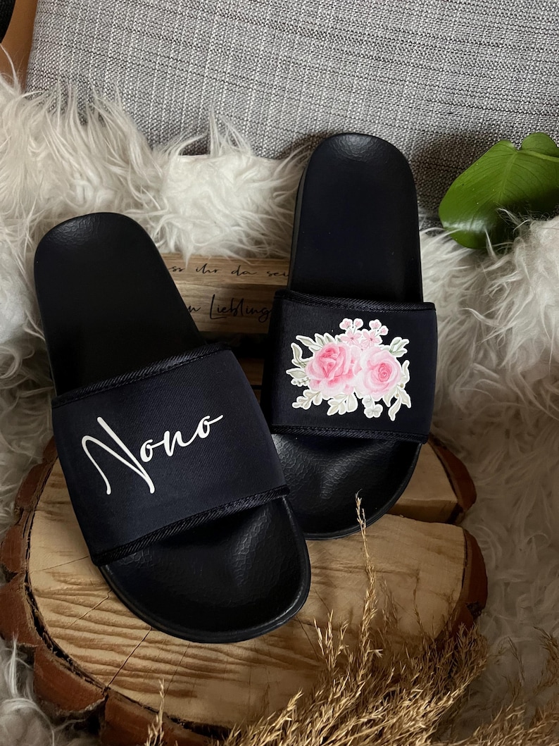 May include: A pair of black slides with a floral design on one slide and the name "Nono" written on the other slide.