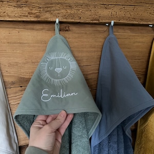 May include: Three hooded towels hanging on hooks. The middle towel is green with a white embroidered lion face and the name "Emilian". The towel on the left is white and the towel on the right is blue.