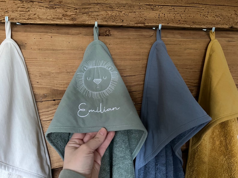 May include: Three hooded towels hanging on hooks. The middle towel is green with a white embroidered lion face and the name "Emilian". The towel on the left is white and the towel on the right is blue.