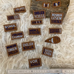 Könnte beinhalten: Eine Sammlung brauner Leder-Patches mit blau gesticktem Text. Die Patches zeigen Sätze wie "KIGA kind", "ENDLICH SCHULE", "LIEBLINGS Erzieher", "MEIN 1. SCHULTAG", "MALEN SINGEN SPIELEN", "VORSCHULE", "MANNCHEN", "RECHNEN LESEN SCHREIBEN", "SCHULKIND", "SCHUL kind", "KINDER GARTEN", "ENDLICH KIGA", "1x1 ABC", "STREBER" und "LIEBLINGS Lehrer".