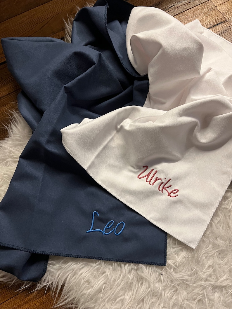 May include: Two personalized towels, one navy blue with the name "Leo" embroidered in blue thread, and one white with the name "Ulrike" embroidered in red thread.