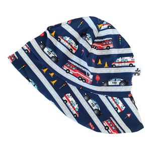 May include: A navy blue bucket hat featuring a road design with white stripes and various emergency vehicles, including fire trucks, ambulances, and police cars. The hat also includes traffic cones, stop signs, and traffic lights. The hat is designed for children.