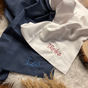 Può includere: Due asciugamani personalizzati, uno blu scuro con il nome "Leo" ricamato in filo blu, e uno bianco con il nome "Ulrike" ricamato in filo rosa.
