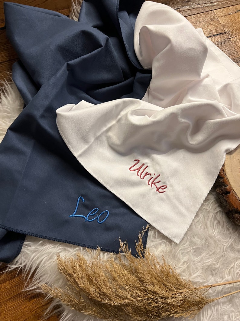 May include: Two personalized towels, one navy blue with the name "Leo" embroidered in blue thread, and one white with the name "Ulrike" embroidered in pink thread.