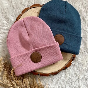 Könnte beinhalten: Zwei Strickmützen mit braunen Bären-Patches. Eine Mütze ist rosa und die andere ist blau.