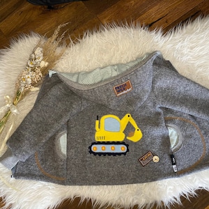 Könnte beinhalten: Graue Kapuzenjacke mit gelbem Bagger-Applikation. Die Jacke hat ein gestreiftes Futter und zwei Taschen. Die Jacke ist aus weichem Material gefertigt und hat einen braunen Aufnäher mit der Aufschrift "FRECH WILD".