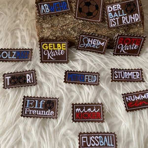 Può includere: Una collezione di toppe in feltro marrone con ricami bianchi, blu, rossi e gialli. Le toppe presentano parole e frasi a tema calcistico, tra cui "Abwehr", "Gelbe Karte", "Tor", "Elf Freunde", "Mittelfeld", "Stürmer", "Der Ball Ist Rund", "Heim Spiel", "Rote Karte", "Nummer Ein", "Mini Kicker" e "Fussball".