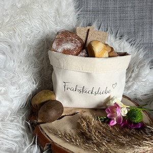 Op de afbeelding: Een witte canvas tas met de tekst "Frühstückliebe" erop gedrukt. De tas is gevuld met broodjes en stokbrood. De tas staat op een houten schijf met een paar halmen gedroogd gras en een paarse bloem.