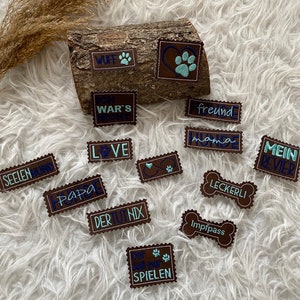 Könnte beinhalten: Eine Sammlung brauner Lederpatches mit verschiedenen Hunde-bezogenen Wörtern und Sätzen, die in blauer und hellblauer Farbe gestickt sind. Die Wörter beinhalten "Wuff", "War's", "Freund", "Love", "Mama", "Seelenhund", "Papa", "Der Nix", "Leckerli", "Mein Revier", "Spiel", und "Impfpass".