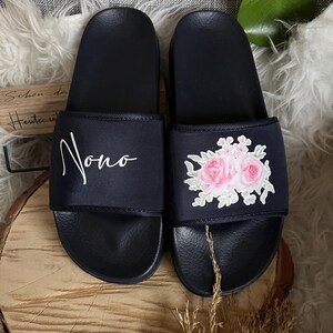 May include: A pair of black slides with a floral design on one slide and the word "Nono" written on the other slide.