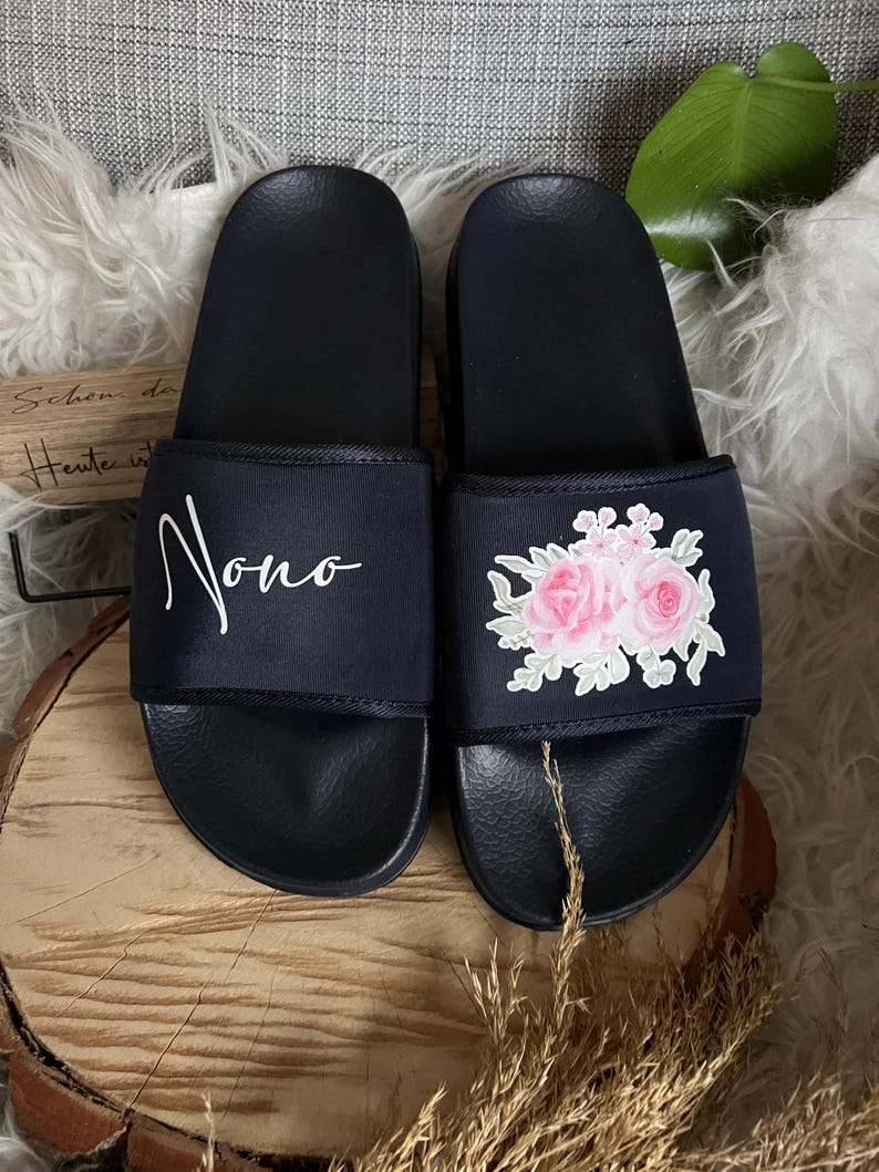May include: A pair of black slides with a floral design on one slide and the word "Nono" written on the other slide.