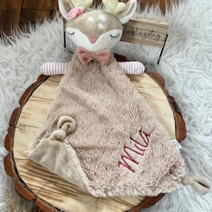 Peut inclure: Une couverture de sécurité en peluche beige avec une tête de cerf attachée. La couverture a un nœud rose et le mot "Mia" brodé dessus. Le cerf a des oreilles roses et une fleur rose sur la tête. Un panneau en bois avec le texte "Lieblingstag" est en arrière-plan.
