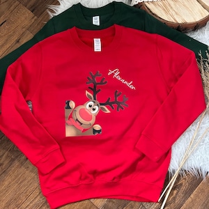 Könnte beinhalten: Ein rotes Sweatshirt mit einem Cartoon-Rentier-Motiv und dem Namen "Alexander" in Schreibschrift. Im Hintergrund ist ein grünes Sweatshirt teilweise zu sehen. Das Rentier hat eine rote Nase und schaut aus der Ecke.