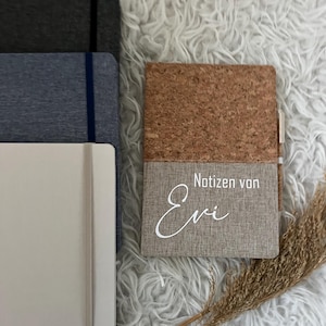 Könnte beinhalten: Eine Auswahl an Notizbüchern in Grau-, Blau- und Weißtönen. Ein Notizbuch hat einen Kork- und Stoffeinband mit dem Text "Notizen von Evi". Ein Stift steckt im Notizbuch. Die Notizbücher liegen auf einer flauschigen, weißen Oberfläche.