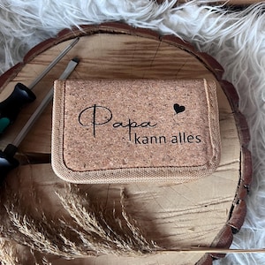 Può includere: Un portafoglio rettangolare in sughero con le parole "Papa kann alles" e un simbolo a cuore stampato sul davanti. Il portafoglio è appoggiato su una superficie di legno con un'estetica naturale e rustica. Sono visibili due cacciaviti.