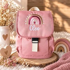 Könnte beinhalten: Ein altrosa Rucksack mit einem gestickten Regenbogenmotiv und einem kleinen Herz in der Mitte. Der Name Cleo ist in weißer Schreibschrift unter dem Regenbogen eingestickt. Der Rucksack hat einen Tragegriff, einen Klappverschluss und eine schwarze Kunststoffschnalle.