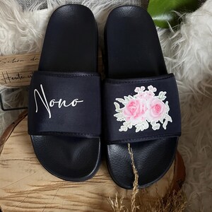 May include: A pair of black slide sandals with a white floral design and the word "Nono" written on one sandal.