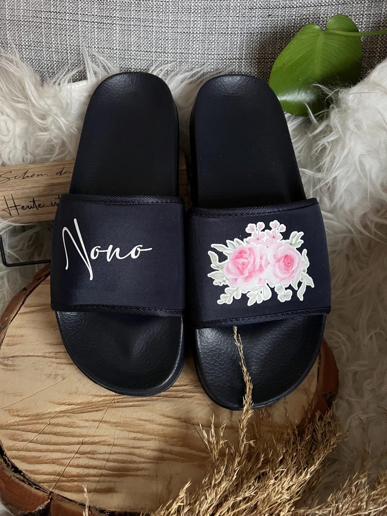 May include: A pair of black slide sandals with a white floral design and the word "Nono" written on one sandal.