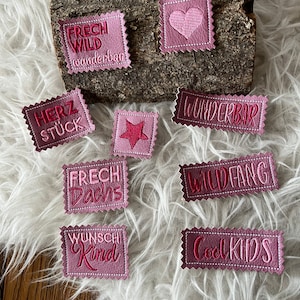 Könnte beinhalten: Neun rosa und rote Filzpatches mit gesticktem Text. Die Patches beinhalten "Frech Wild", "Wunderbar", "Herz Stuck", "Frech Dachs", "Wildfang", "Cool Kids", "Wunschi Kind" und zwei Patches mit einem Herz und einem Stern.