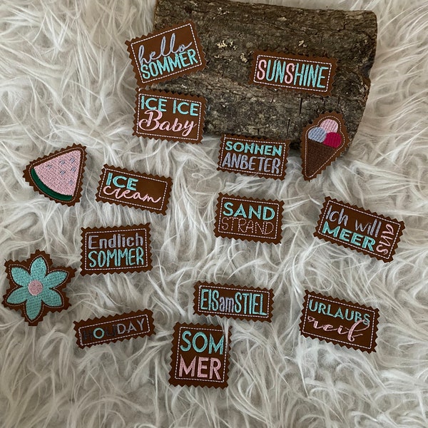May include: A collection of brown leather patches with various summer-themed designs and text, including "hello sommer", "ice ice baby", "sunshine", "sonnen anbeter", "sand strand", "ich will meers", "endlich sommer", "eisamstiel", "urlaubs reif", "o day", and "som mer".