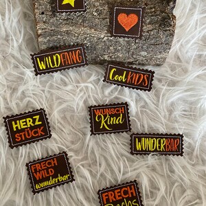 Könnte beinhalten: Eine Auswahl an braunen Stoffaufnähern mit gestickten Designs und Text. Die Aufnäher zeigen einen gelben Stern, ein orangefarbenes Herz und Sätze wie "WILD FANG" und "COOL KIDS". Die Aufnäher sind auf einem Holzstück angeordnet.