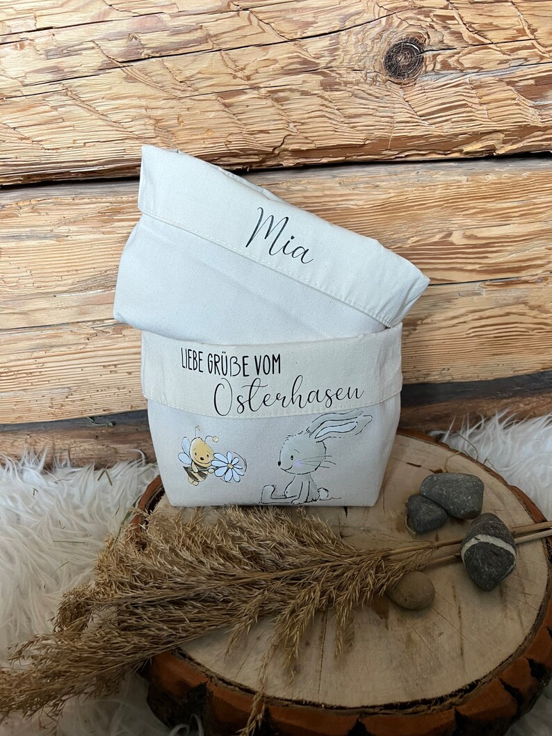 K&ouml;nnte beinhalten: Ein wei&szlig;er Canvas-Osterkorb mit dem Text "Liebe Gr&uuml;&szlig;e vom Osterhasen" und einer kleinen Illustration eines Hasen und einer Biene. Der Korb ist oben umgeklappt und tr&auml;gt den Namen "Mia" aufgeschrieben.