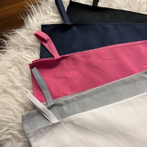 May include: Five microfiber towels in different colors, stacked on top of each other. The towels are white, gray, pink, navy blue, and black. Each towel has a loop at the top for hanging.