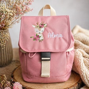 Può includere: Uno zaino rosa con chiusura a patta e cinturino color crema. La patta presenta un cervo ricamato con una corona floreale e il nome "Alma" in corsivo bianco. Lo zaino è appoggiato su una superficie di legno.