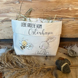 K&ouml;nnte beinhalten: Ein wei&szlig;er Stoff-Osterkorb mit einer Bienen- und Hasenillustration. Der Korb hat den Text "Liebe Gr&uuml;&szlig;e vom Osterhasen" aufgedruckt.