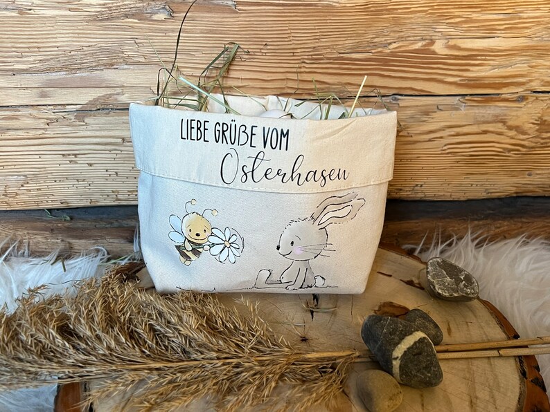 K&ouml;nnte beinhalten: Ein wei&szlig;er Stoff-Osterkorb mit einer Bienen- und Hasenillustration. Der Korb hat den Text "Liebe Gr&uuml;&szlig;e vom Osterhasen" aufgedruckt.
