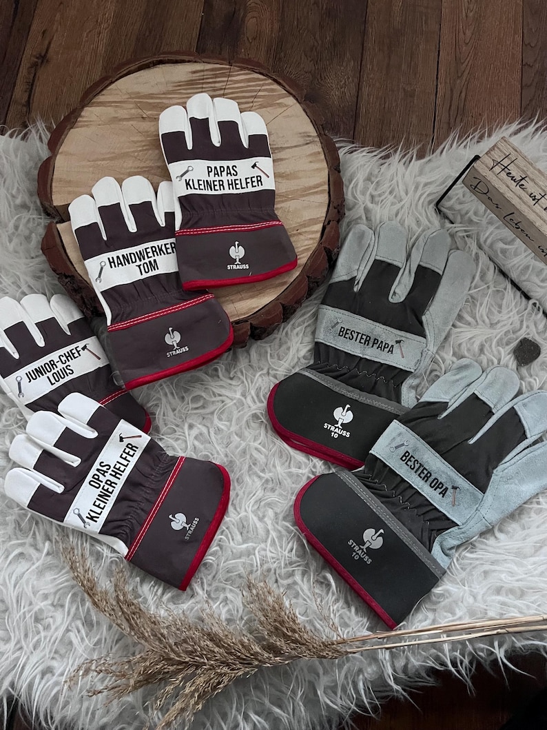 Może przedstawiać: Kolekcja rękawic roboczych w r&oacute;żnych kolorach, w tym białym, szarym i brązowym. Każda rękawica zawiera napisy takie jak "Handwerker Tom" i "Bester Papa". Rękawice mają czerwone wykończenia i logo "Strauss".