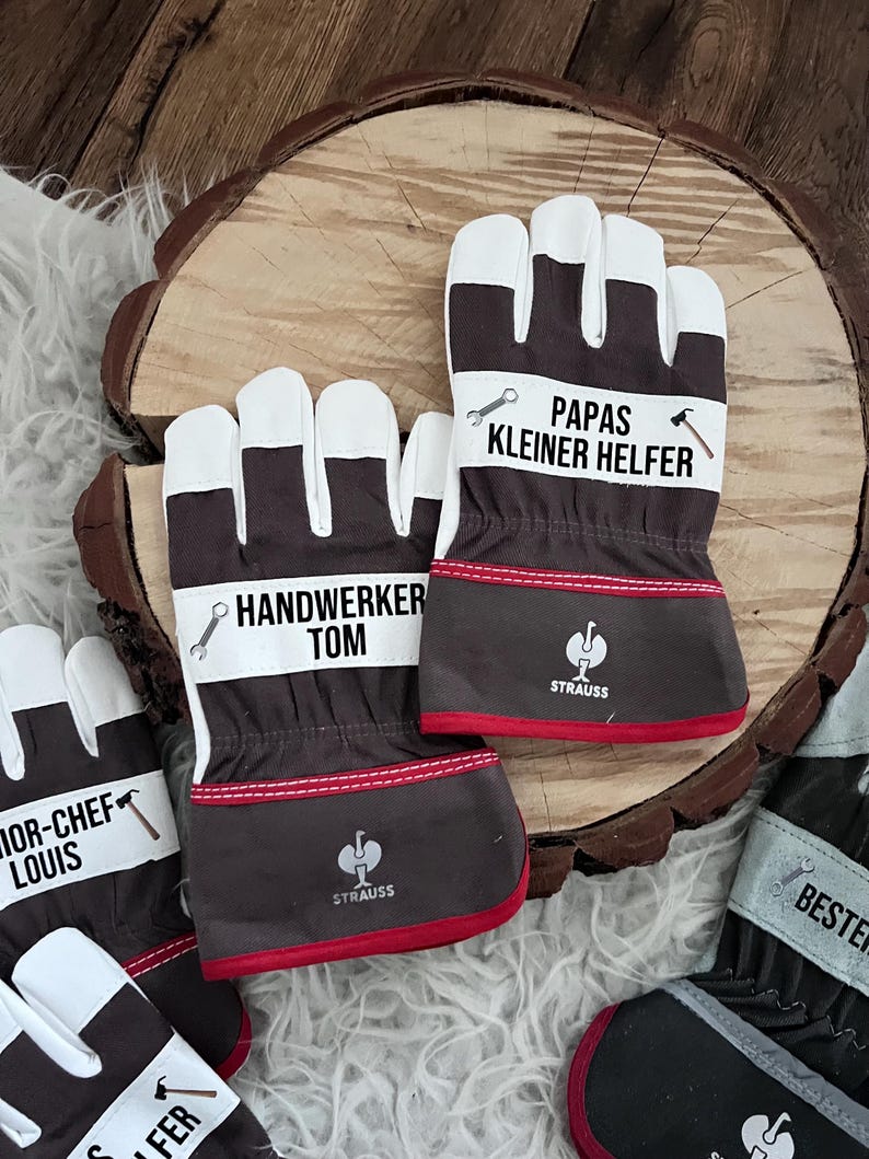 Może przedstawiać: Brązowo-białe rękawice robocze z czerwonymi wykończeniami, z napisami takimi jak "HANDWERKER TOM" i "PAPAS KLEINER HELFER". Rękawice mają grafikę młotka i klucza. Rękawice leżą na drewnianej powierzchni.