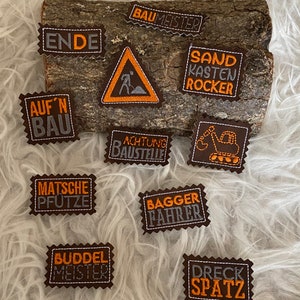 May include: A set of ten embroidered iron-on patches with construction-themed designs. The patches are brown with orange and gray embroidery. The patches feature phrases like "Sand Kasten Rocker", "Buddel Meister", and "Dreck Spatz".