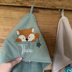 Könnte beinhalten: Ein grünes Kapuzenhandtuch mit einem Fuchs, der über den Rand schaut, und dem Namen "Theo", der auf der Vorderseite gestickt ist. Das Handtuch hängt an einer Holzoberfläche.