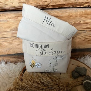 K&ouml;nnte beinhalten: Eine wei&szlig;e Stofftasche mit einer Bienen- und Hasenillustration. Die Tasche hat einen wei&szlig;en Stoffdeckel mit dem Namen "Mia" darauf geschrieben. Die Tasche hat auch ein wei&szlig;es Stoffband mit dem Text "Liebe Gr&uuml;&szlig;e vom Osterhasen".