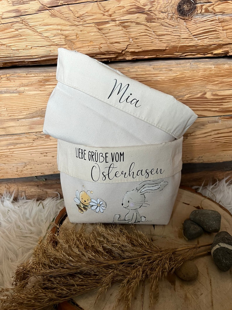 K&ouml;nnte beinhalten: Eine wei&szlig;e Stofftasche mit einer Bienen- und Hasenillustration. Die Tasche hat einen wei&szlig;en Stoffdeckel mit dem Namen "Mia" darauf geschrieben. Die Tasche hat auch ein wei&szlig;es Stoffband mit dem Text "Liebe Gr&uuml;&szlig;e vom Osterhasen".