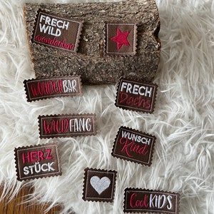 May include: A collection of brown embroidered patches with white and pink text and designs, including a star and a heart. The patches are arranged on a piece of wood and a fluffy white surface.
